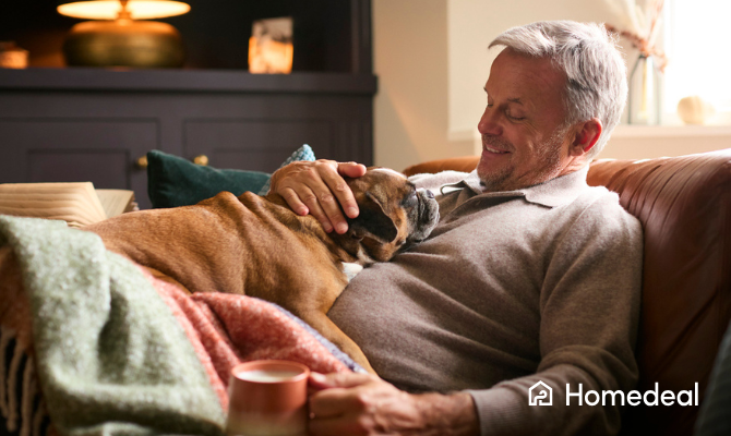 Man op leeftijd ligt gezellig met hond en kop koffie onder een kleedje op de bank