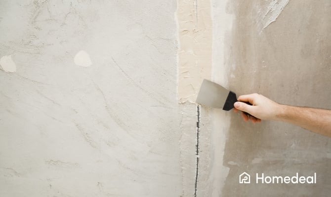 Een hand brengt met een plamuurmes vulmiddel aan over een met wapeningsband versterkte scheur in een muur om het oppervlak glad te maken.