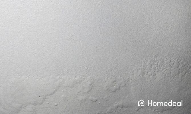 Witte muur met schade door optrekkend vocht, zichtbaar als vochtplekken en afbladderende verf aan de onderzijde van de muur.