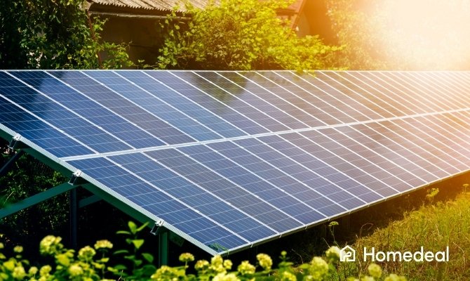 Rij zonnepanelen op een metalen frame gemonteerd op de grond, omringd door groene beplanting en met een woning op de achtergrond in zonlicht.