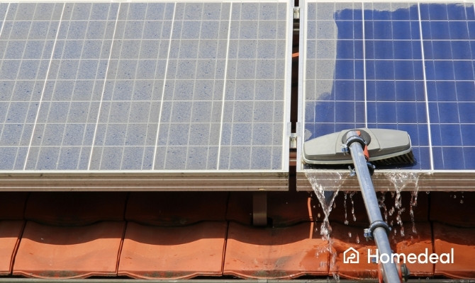 Close-up van het schoonmaken van zonnepanelen op een rood pannendak met een waterdoorlatende borstel, waarbij water langs de panelen stroomt.