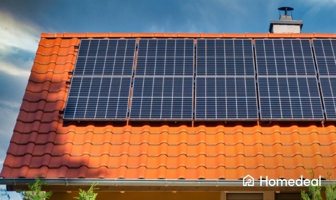 Monokristallijne zonnepanelen op rood pannendak van een huis met blauwe lucht.