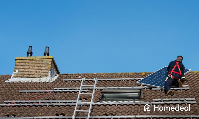 Zonnepanelen installateur met veiligheidsharnas tilt een zonnepaneel op het dak van een woning om het te monteren op een schuin pannendak met bevestigingsrails.
