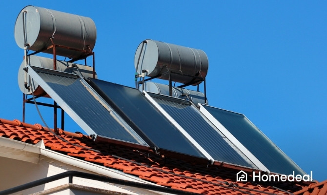Meerdere vlakke plaat zonnecollectoren met opslagtanks op een pannendak, ontworpen voor het verwarmen van water met zonne-energie, onder een felblauwe lucht.