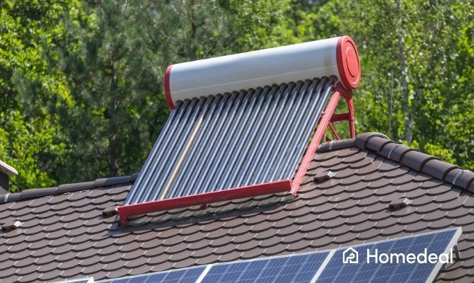 Zonneboiler met vacuümbuizen en rood frame geïnstalleerd op een schuin pannendak, naast traditionele zonnepanelen, met groene bomen op de achtergrond.