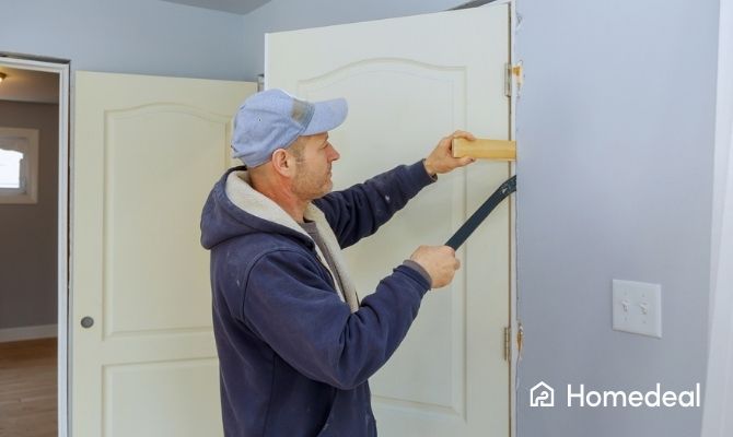 Man die met een koevoet en houten blok een binnendeur en kozijn losmaakt tijdens renovatiewerkzaamheden, in een kamer met lichtgrijze muren.