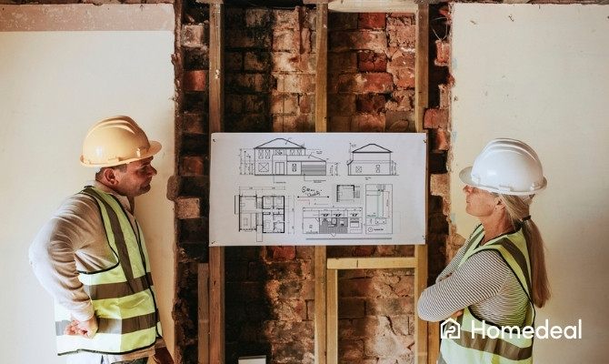 Twee bouwprofessionals met helmen en fluohesjes staan voor een bakstenen muur op een renovatiewerf en bestuderen een opgehangen bouwtekening met voor- en bovenaanzichten van een woning.