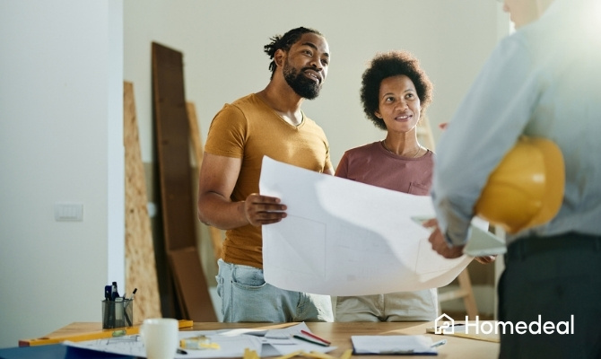 Man en vrouw staan samen met een architect in een ruimte en houden een grote bouwtekening vast. Ze bespreken de plannen voor hun woning, terwijl op de tafel bouwmaterialen, documenten en een helm liggen.