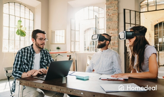 Twee klanten met VR-brillen op krijgen een virtuele rondleiding van een architect aan tafel, terwijl hij uitleg geeft via zijn laptop in een modern, licht kantoor.