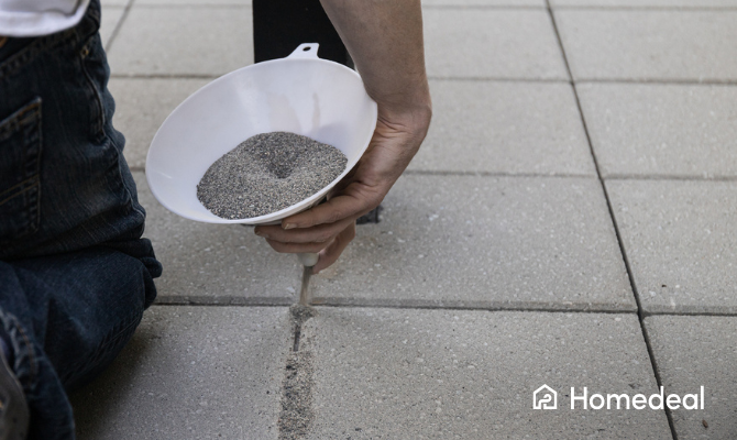 Man zit op zijn knieën en laat voegzand vanuit een trechter tussen de stenen van een nieuw terras lopen