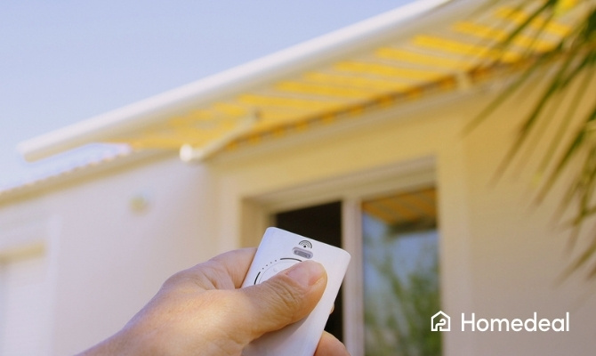 Hand die een afstandsbediening vasthoudt om een elektrisch zonnescherm bij een woning te bedienen.