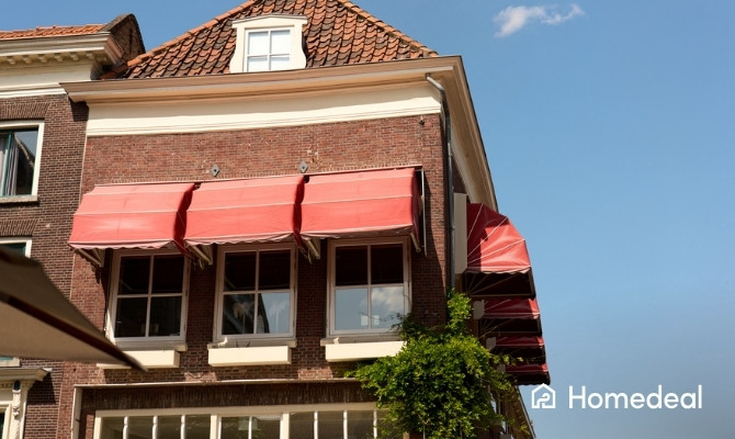 Gevel van een klassiek Nederlands bakstenen pand met rode knikarmschermen boven de ramen op de eerste verdieping, gefotografeerd van onderaf tegen een blauwe lucht.