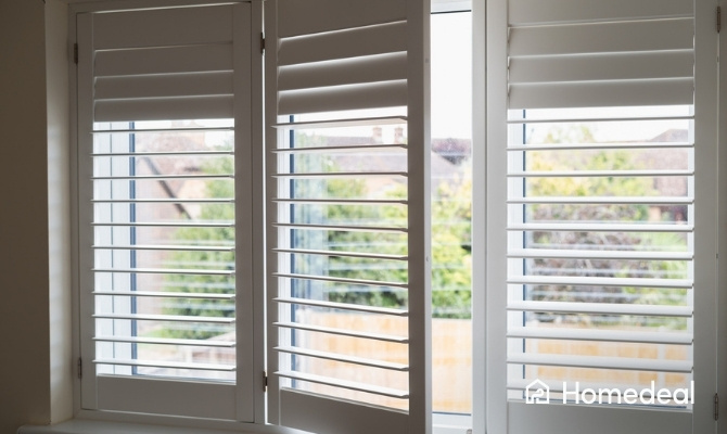 Witte shutters met verstelbare lamellen voor een raam, waarvan één paneel deels openstaat en uitzicht biedt op de tuin.