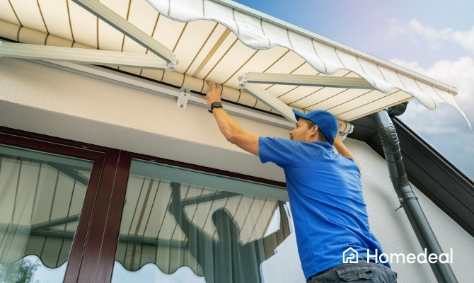 Monteur in blauw shirt en pet plaatst een creme-kleurige uitvalscherm aan de gevel van een woonhuis boven een schuifpui.
