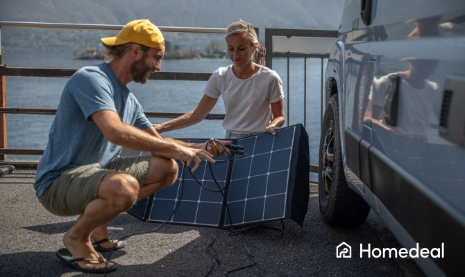 Man en vrouw sluiten samen een draagbaar zonnepaneel aan bij hun camper, geparkeerd aan het water met een berglandschap op de achtergrond.