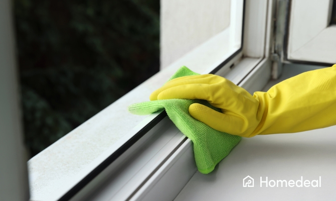 Hand met gele schoonmaakhandschoen maakt een wit kunststof kozijn schoon met een groene microvezeldoek tijdens kunststof kozijnen onderhoud en schoonmaken.