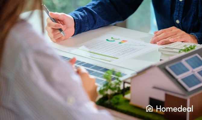 Twee personen bespreken het vermogen van zonnepanelen aan de hand van een energieprestatie-overzicht, naast een maquette van een huis met zonnepanelen op het dak.