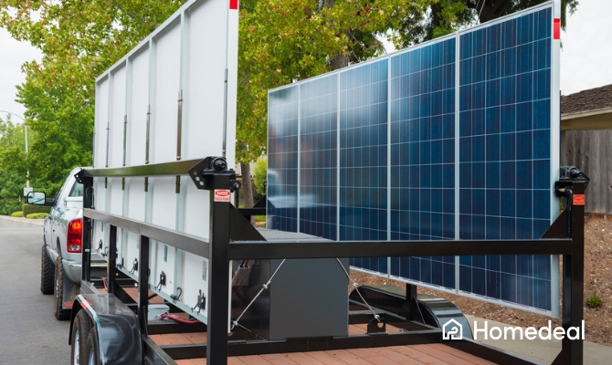 Een aanhanger met gemonteerde zonnepanelen wordt voortgetrokken door een pickuptruck in een woonwijk, bedoeld voor mobiele stroomopwekking of tijdelijke energievoorziening.
