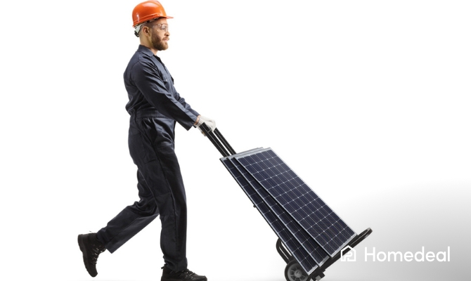 Een man in werkpak en veiligheidshelm verplaatst meerdere zonnepanelen op een steekwagen tegen een witte achtergrond, klaar voor installatie.