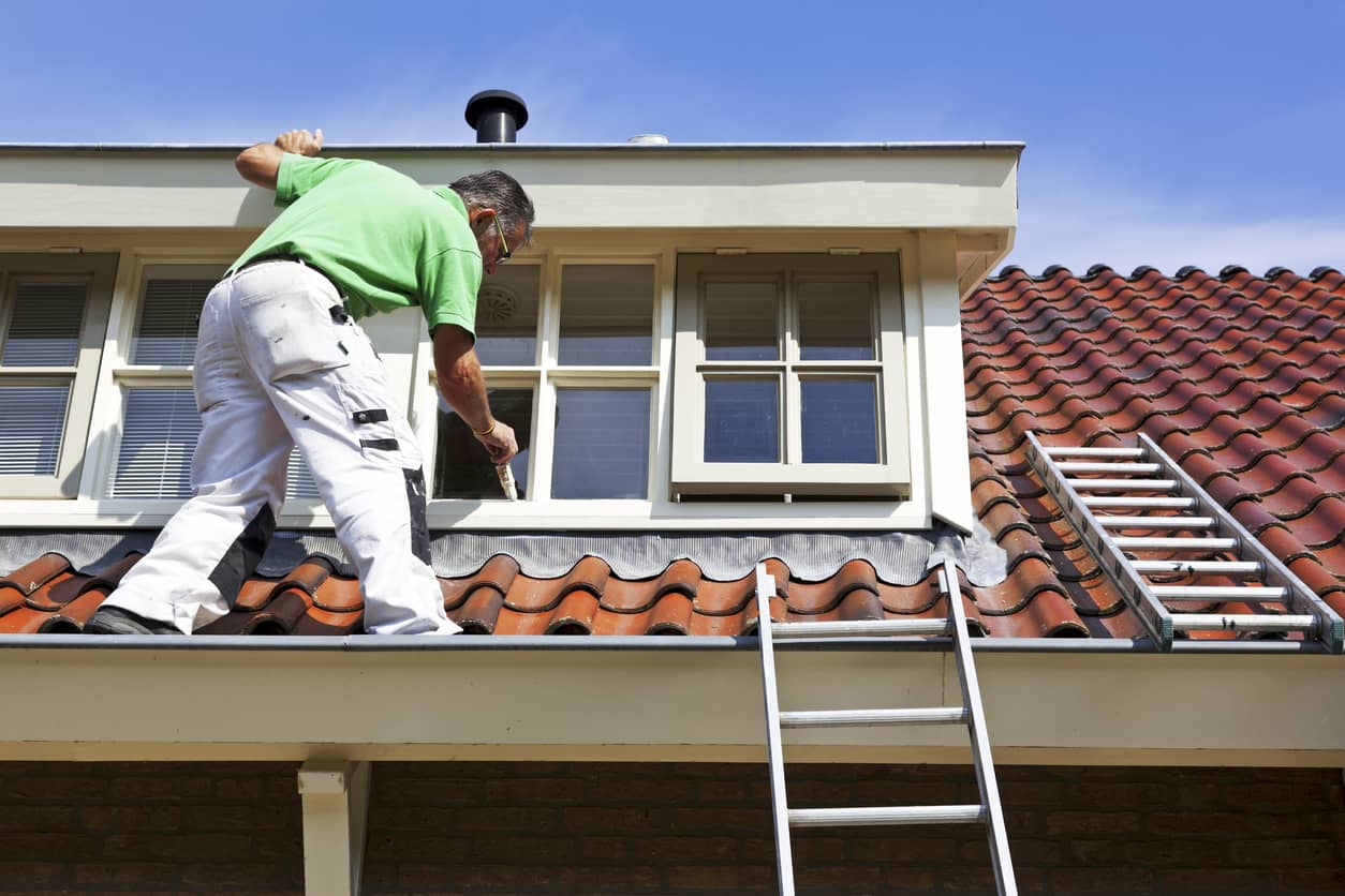 Schilder op dak die een dakkapel verft met kwast