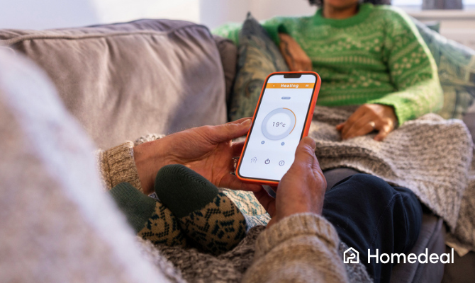 Man en vrouw zitten onder een dekentje op de bank, man past op zijn telefoon in een app de verwarmingstemperatuur aan